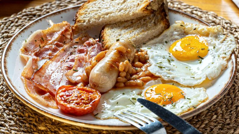 A hyper-realistic, high-angle shot of a classic Full English breakfast on a white ceramic plate, resting on a rustic wooden table. The lighting is warm morning sunlight. Surrounding the plate are faint, translucent logistical map lines and small icons (ships, trucks, wheat sheaves) connecting the ingredients to a blurred background of a British rolling countryside and a distant container ship, symbolizing the local and global journey.