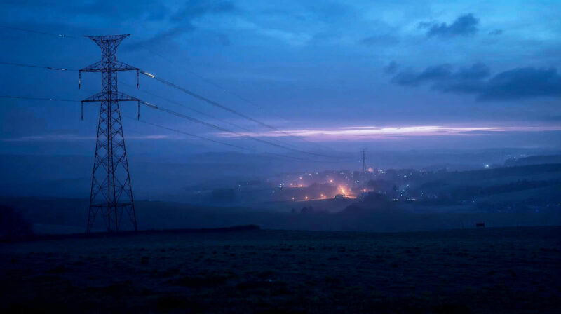 A hyper-realistic, cinematic wide shot of a misty British landscape at dusk. In the foreground, a classic steel pylon stands tall against a deep blue and purple sky, with faint glowing lines of energy visualized flowing along the wires. In the distance, the warm amber glow of a small British village nestles in the rolling green hills. The style should be atmospheric and documentary-like, evoking a sense of quiet power and connection, similar to a BBC high-end documentary intro.