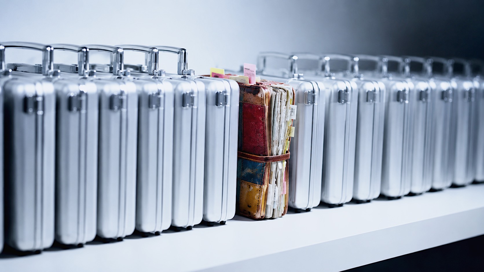 A long row of identical, perfectly polished, sleek silver briefcases sitting on a minimalist white table. The briefcases represent flawless AI business writing. Right in the middle of the row, breaking the pattern, is a single, brightly coloured, slightly worn, handmade leather sketchbook overflowing with notes and post-its. Cinematic lighting, shallow depth of field focusing on the colourful sketchbook, 16:9 aspect ratio, high contrast between the identical perfection of the briefcases and the human uniqueness of the sketchbook.